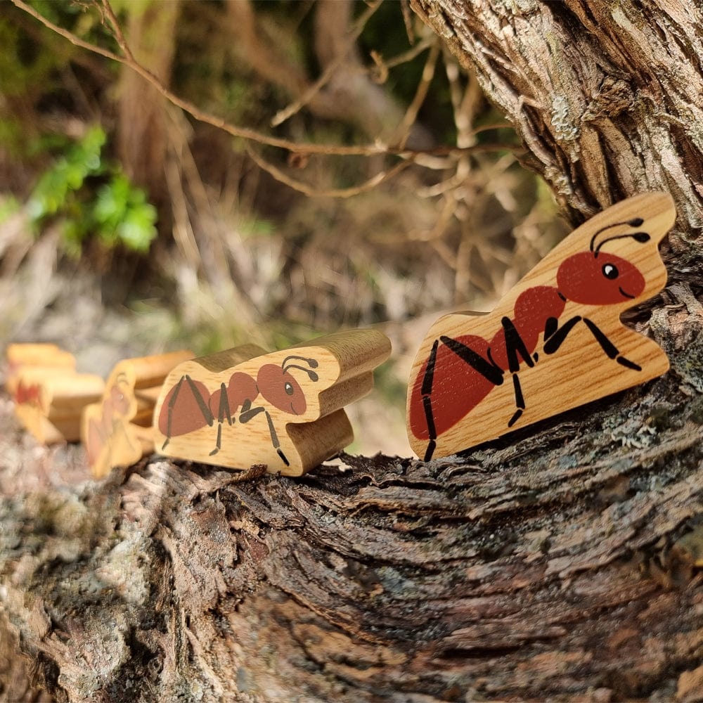 three wooden toy figures of an ant with a natural wood grain edge and red and black colouring. The ants are on a log in an outdoor setting.